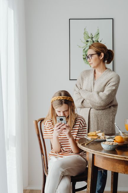 A mother with crossed arms stands beside a daughter using a smartphone at breakfast.